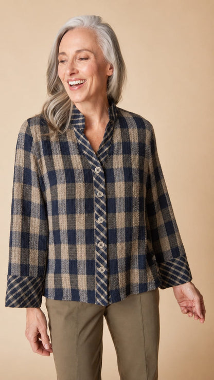 A smiling woman with gray hair wearing a navy and beige plaid button-up shirt and olive pants, standing against a solid beige background.