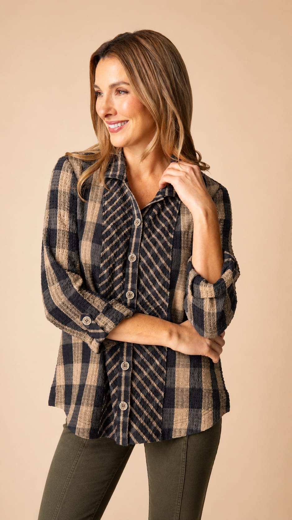 A woman modeling a long-sleeve button-up plaid shirt in navy and beige, paired with dark olive pants, against a solid beige studio background.