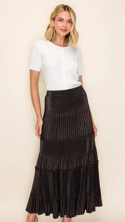 A woman modeling a white short-sleeve top and a black pleated tiered midi skirt against a plain beige studio background.