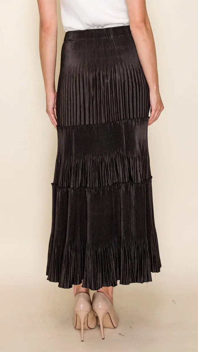 A black pleated tiered maxi skirt worn by a model, paired with a white top and beige high heels, set against a plain beige background.