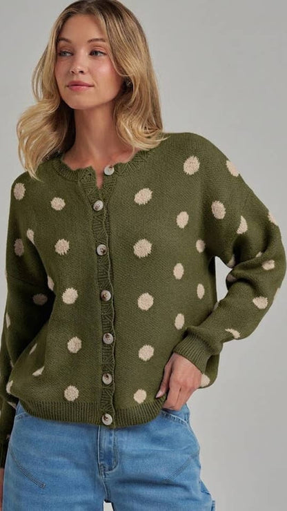 A woman wearing an olive green knit cardigan with beige polka dots and light blue jeans, photographed against a plain light gray studio background.