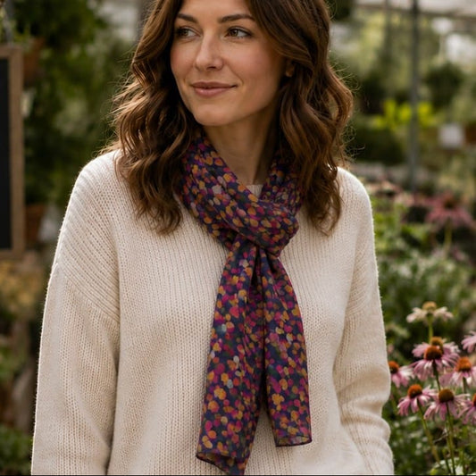 A woman with wavy brown hair wearing a cream knit sweater and a dark purple silk scarf with a small multicolor floral pattern, standing in a garden setting.