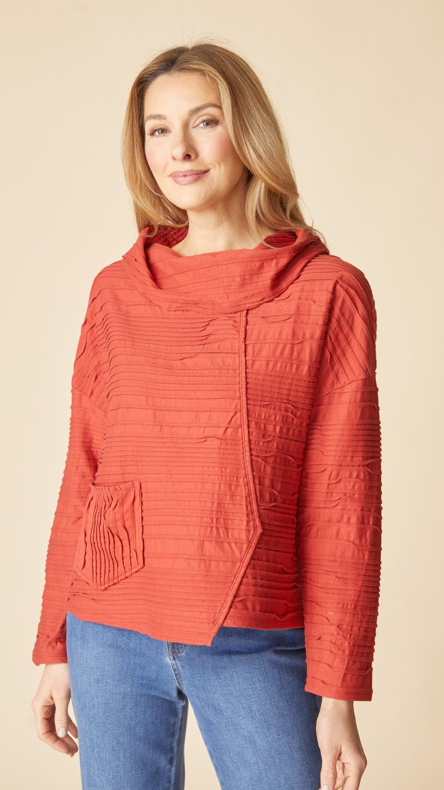 A woman wearing a textured, rust-orange hooded jacket with a cowl neck and front patch pocket, paired with blue jeans, against a solid beige background.