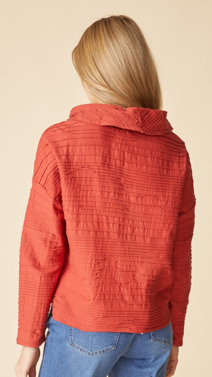 A woman viewed from behind wearing a textured, ribbed orange-red cropped jacket with a high collar and blue jeans, set against a plain beige background.