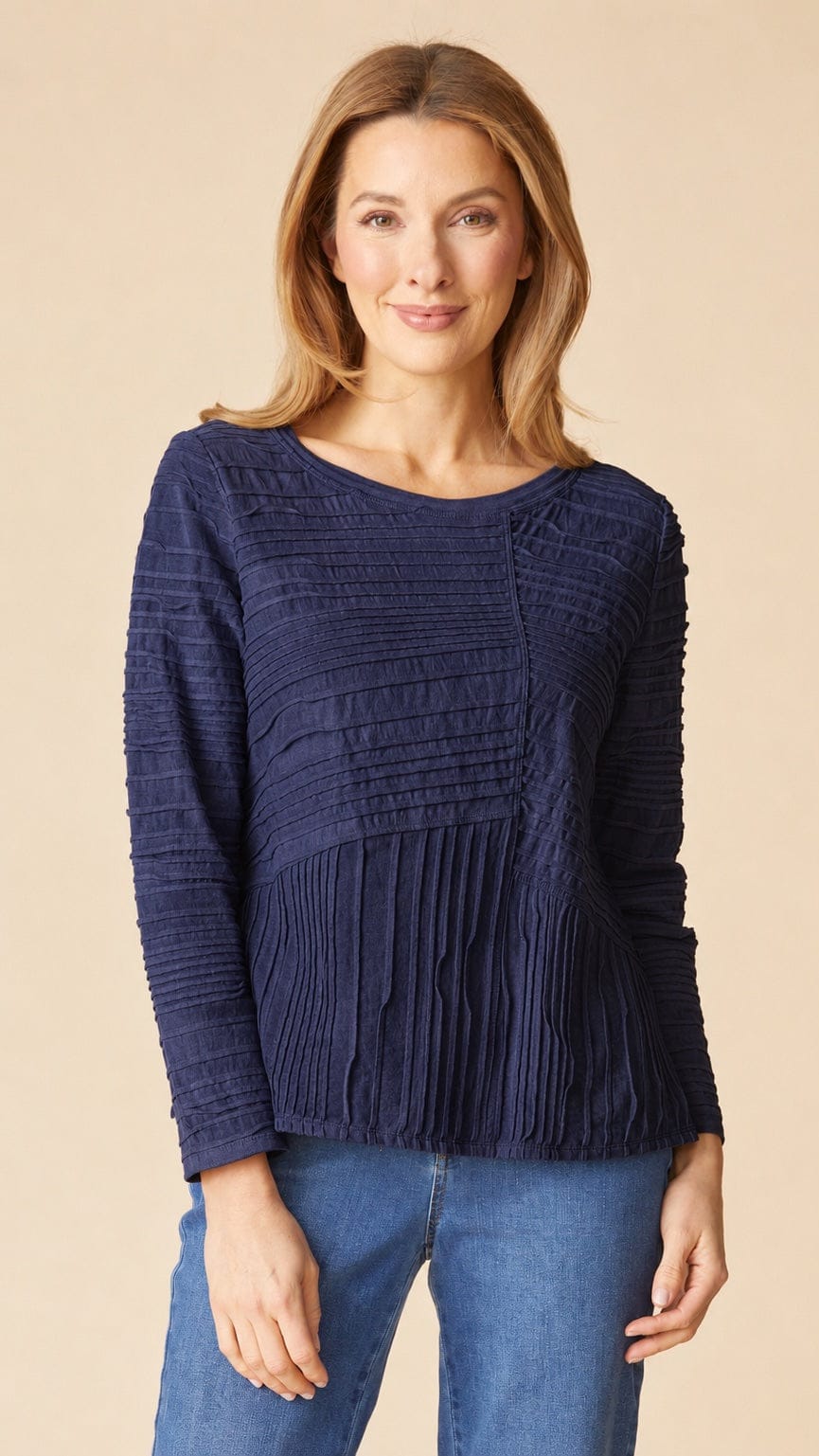 A woman wearing a navy blue long-sleeve knit top with textured horizontal pleating and a gathered peplum hem, paired with blue jeans, against a solid beige background.