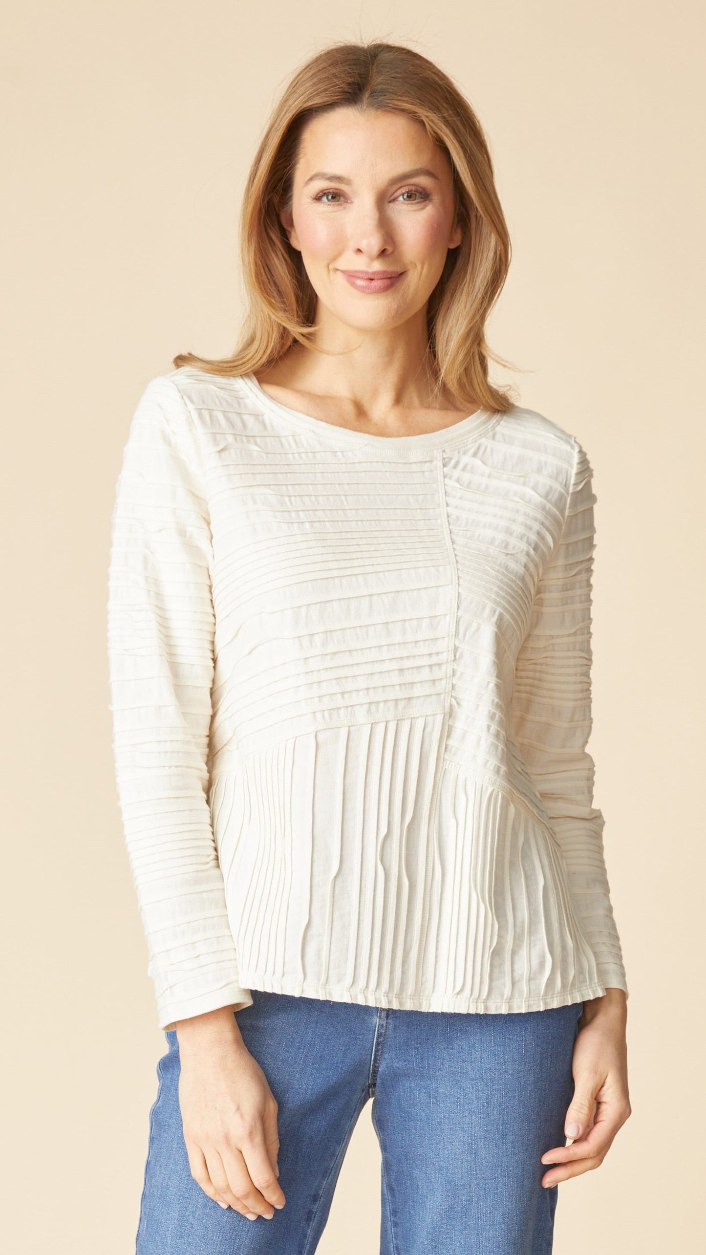 A woman wearing a cream-colored, long-sleeve textured knit sweater with pleated details, paired with blue jeans, against a solid beige background.