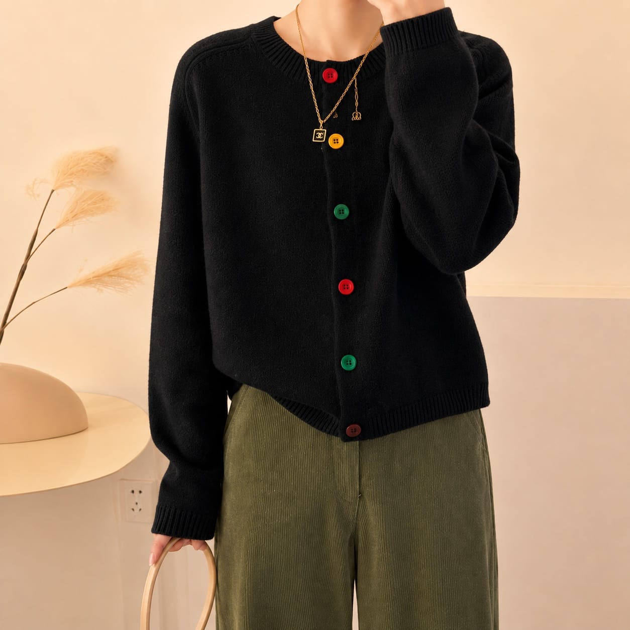 A person wearing a black knit cardigan with colorful buttons in red, yellow, green, and brown, paired with olive green corduroy pants, photographed in a warm indoor setting.