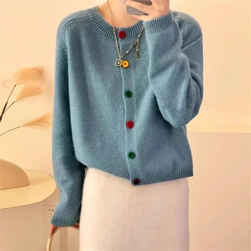 A person wearing a light blue knit cardigan sweater with colorful buttons in red, green, and dark brown, paired with a white skirt. The photo is taken in a mirror selfie style in a warm, beige-toned indoor setting.