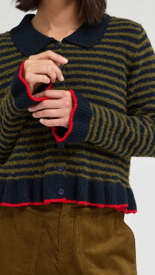 A person wearing a striped knit cardigan with a dark navy collar, cuffs, and hem, accented with bright red trim, paired with brown corduroy pants against a light gray studio background.
