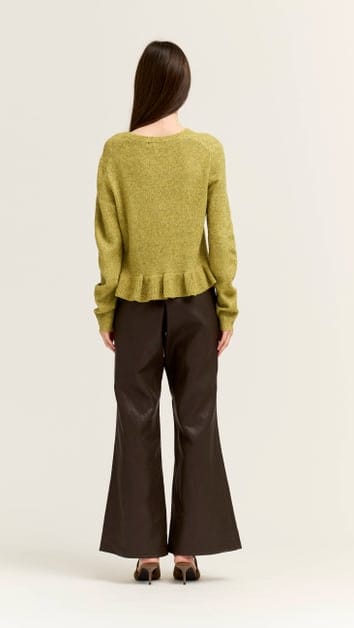 A woman shown from the back wearing a textured olive green long-sleeve sweater with a ruffled hem and dark brown flared pants, standing against a plain light background.