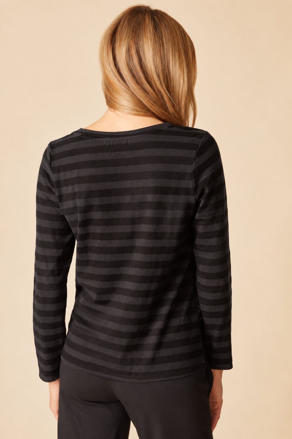 A woman viewed from the back wearing a black long-sleeve top with horizontal dark gray stripes, set against a solid beige background.