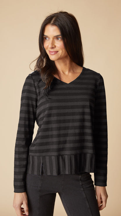 A woman modeling a black and dark gray horizontally striped long-sleeve top with a V-neck and a ruffled hem, paired with black pants, against a solid beige background.