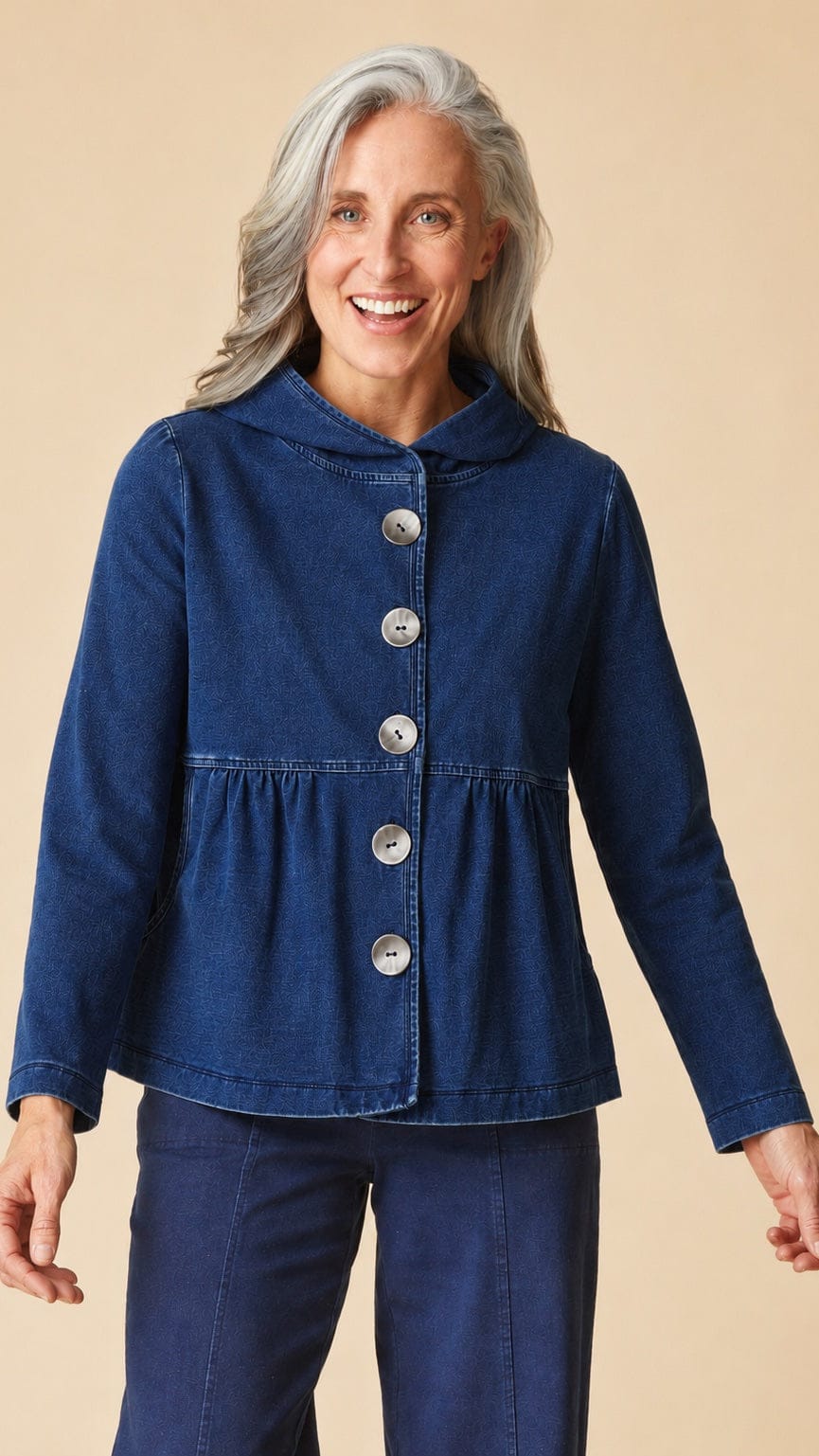 A smiling older woman with gray hair wearing a dark blue hooded button-front jacket with a gathered peplum waist and matching dark blue pants, posed against a solid beige studio background.