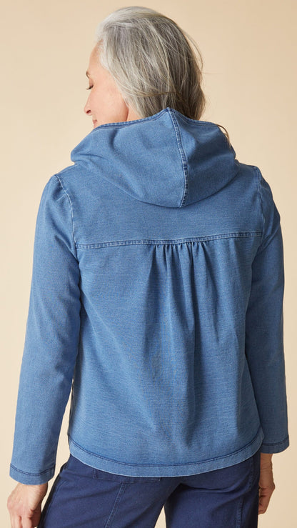 A woman shown from the back wearing a blue hooded jacket with a gathered back seam, paired with dark blue pants, against a solid beige background.