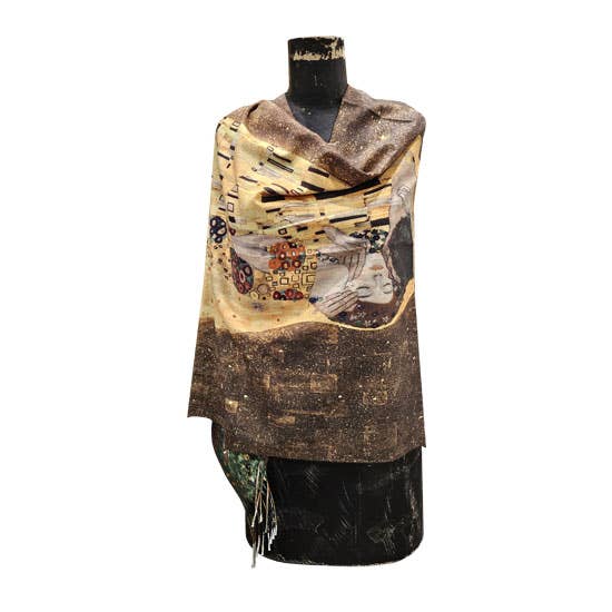A draped shawl or wrap displayed on a black mannequin torso against a plain white background. The shawl has an abstract art print with a mix of brown, beige, black, and gold tones, featuring a stylized face and geometric patterns.