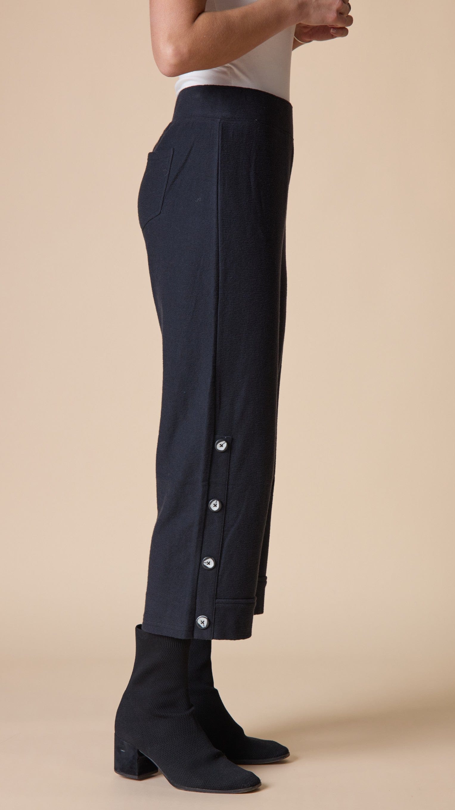 A model is shown from the waist down wearing dark navy blue cropped trousers with decorative silver buttons down the side and black ankle boots, set against a solid beige background.