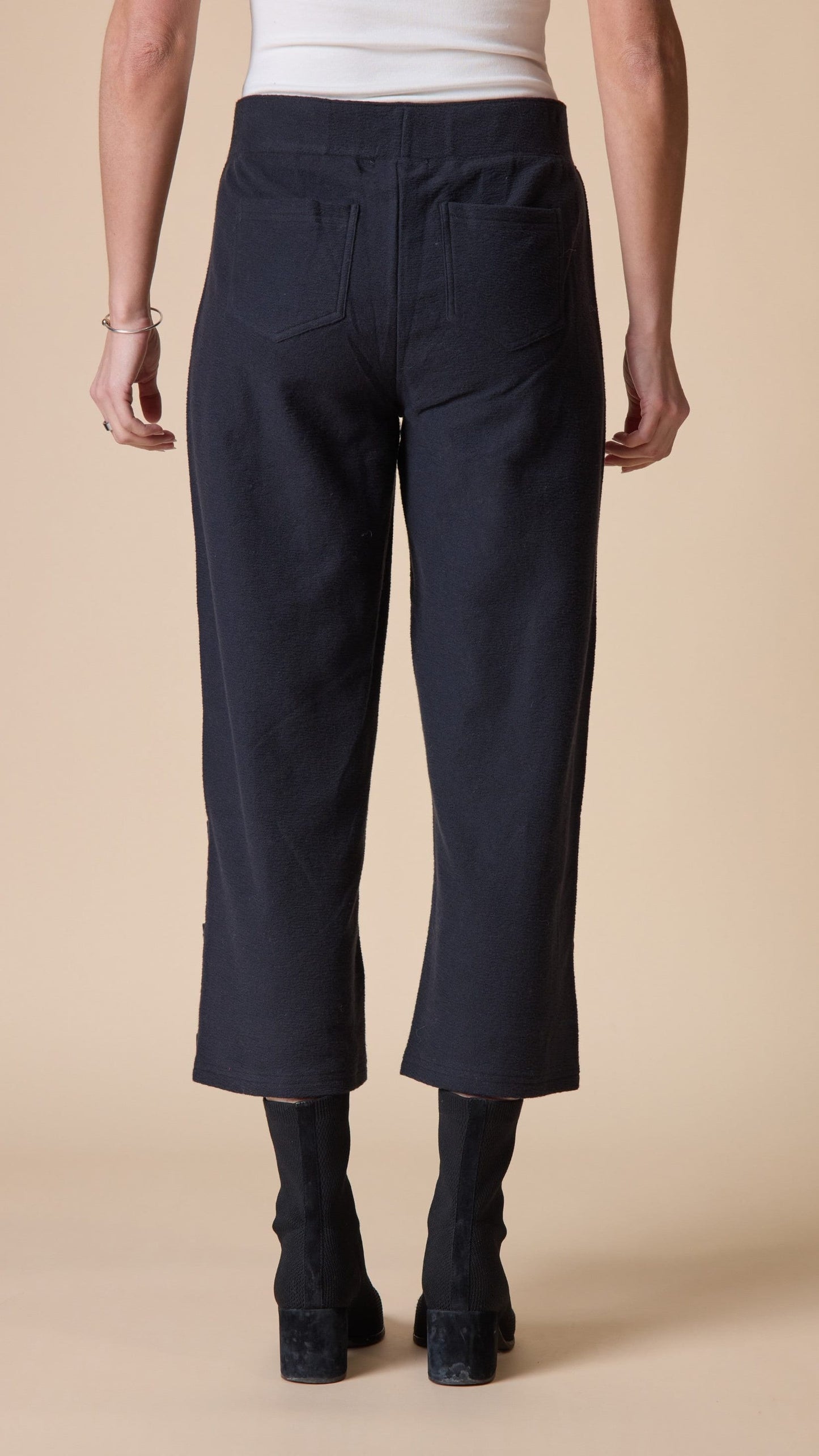 A model shown from the back wearing dark navy cropped pants, a white top, and black ankle boots against a solid beige studio background.