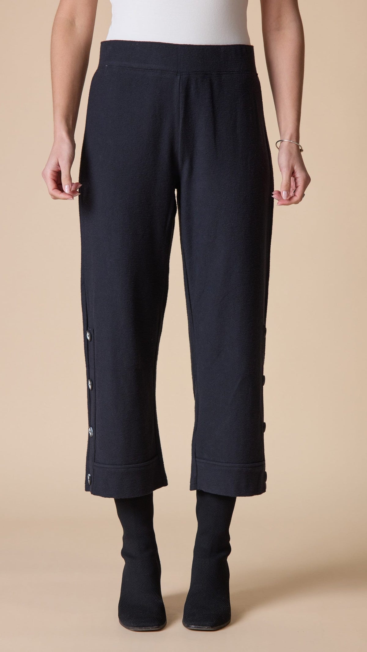 A model is shown from the waist down wearing dark navy cropped pants with button details on the side, paired with black socks and black boots, against a solid beige background.