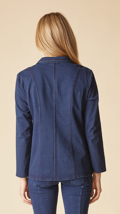 A woman viewed from the back wearing a dark blue denim blazer and matching blue jeans against a solid beige background.