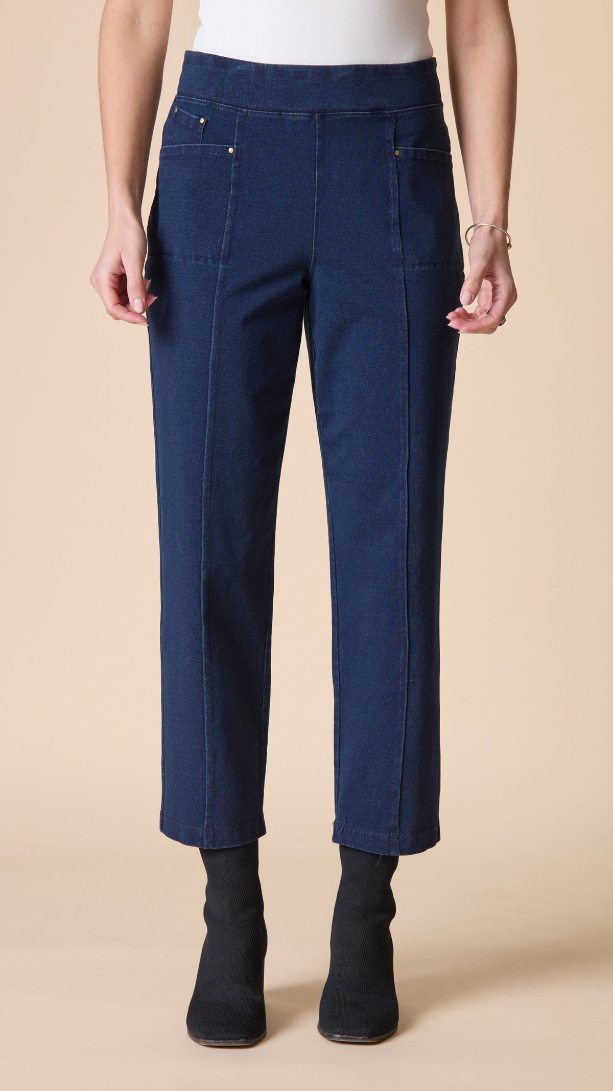 A model wearing dark blue cropped jeans with front patch pockets, paired with black ankle boots, shown from the waist down against a solid beige background.