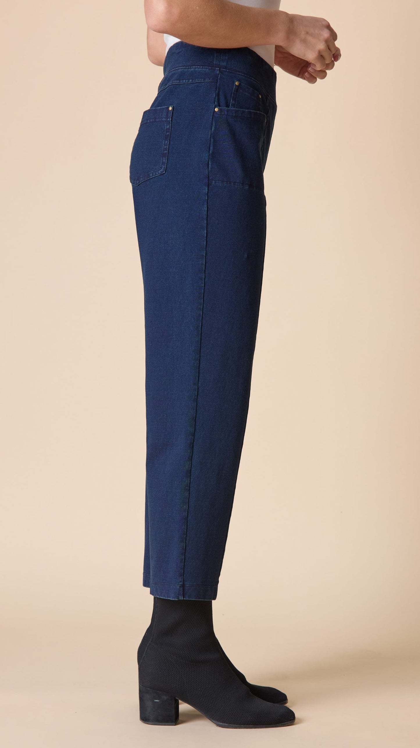 A side-profile view of a person wearing dark blue cropped jeans and black ankle boots against a solid beige studio background.