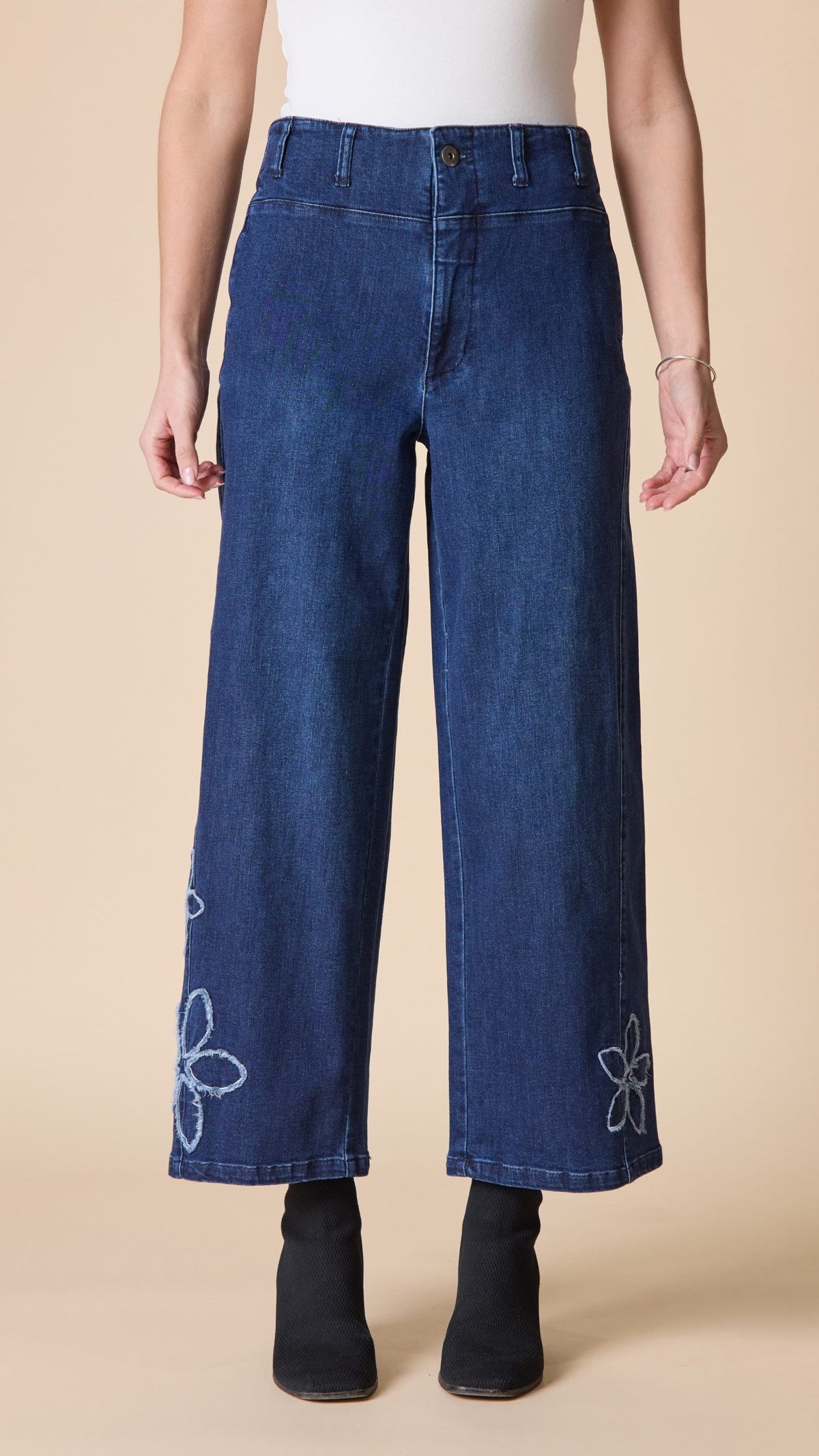 A model is shown from the waist down wearing dark blue wide-leg cropped jeans with white embroidered flower details near the hems, paired with a white top and black ankle boots, against a solid beige background.