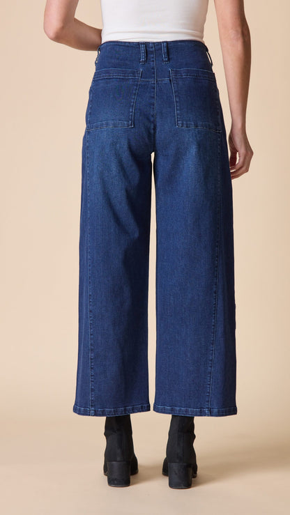 Rear view of a model wearing high-waisted, wide-leg blue denim jeans with a white ribbed top and black ankle boots, set against a solid beige background.