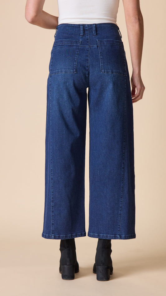 Rear view of a model wearing high-waisted, wide-leg blue denim jeans with a white ribbed top and black ankle boots, set against a solid beige background.