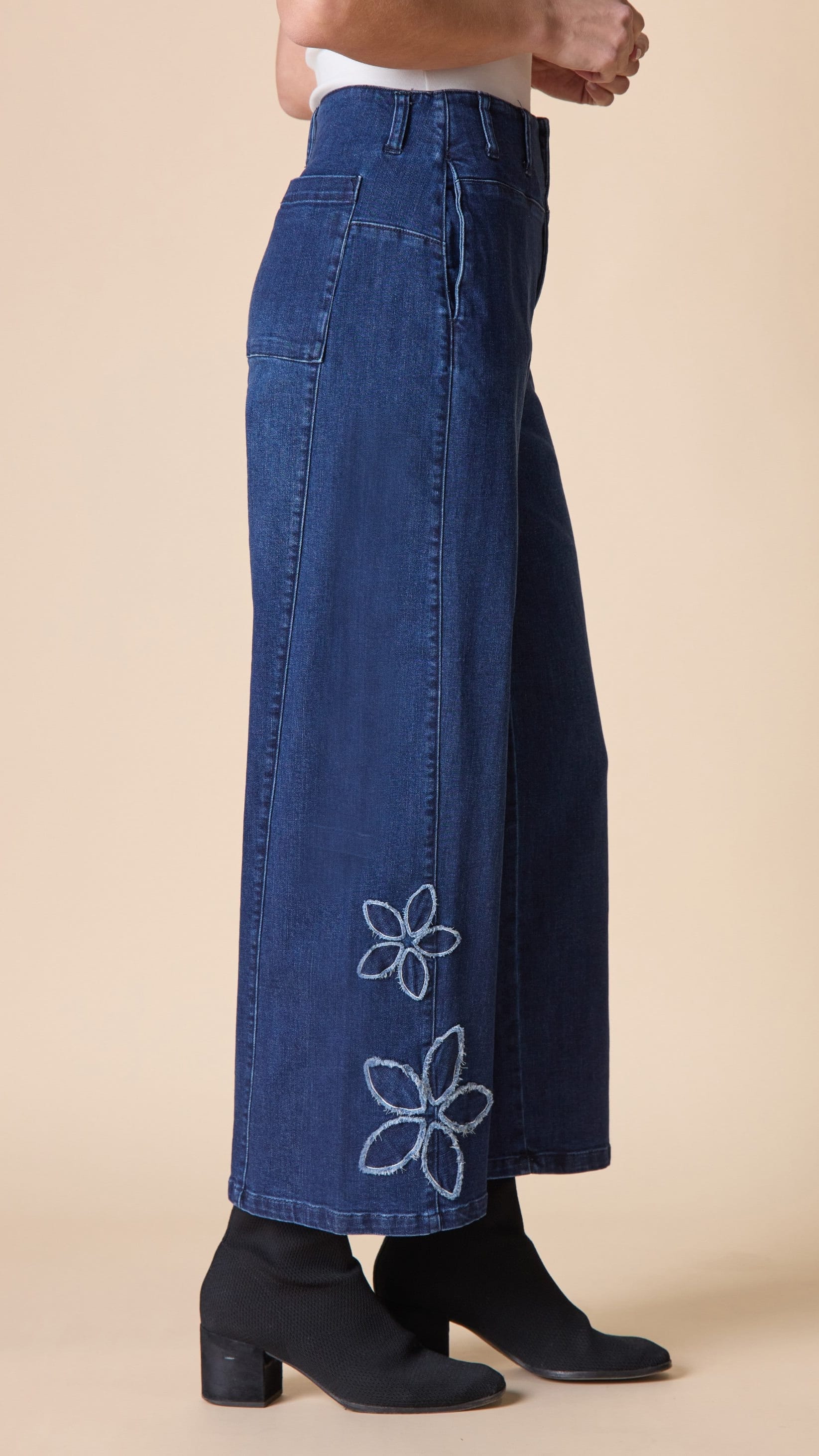 A person wearing dark blue wide-leg denim pants with white embroidered flower motifs near the hem, paired with black ankle boots against a plain beige studio background.