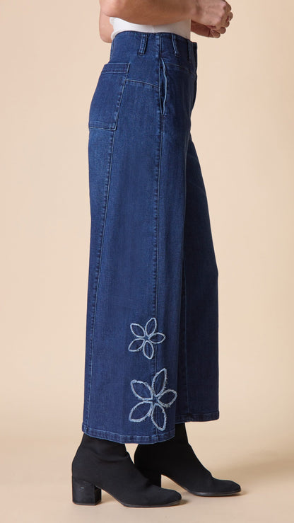 A person wearing dark blue wide-leg denim pants with white embroidered flower motifs near the hem, paired with black ankle boots against a plain beige studio background.