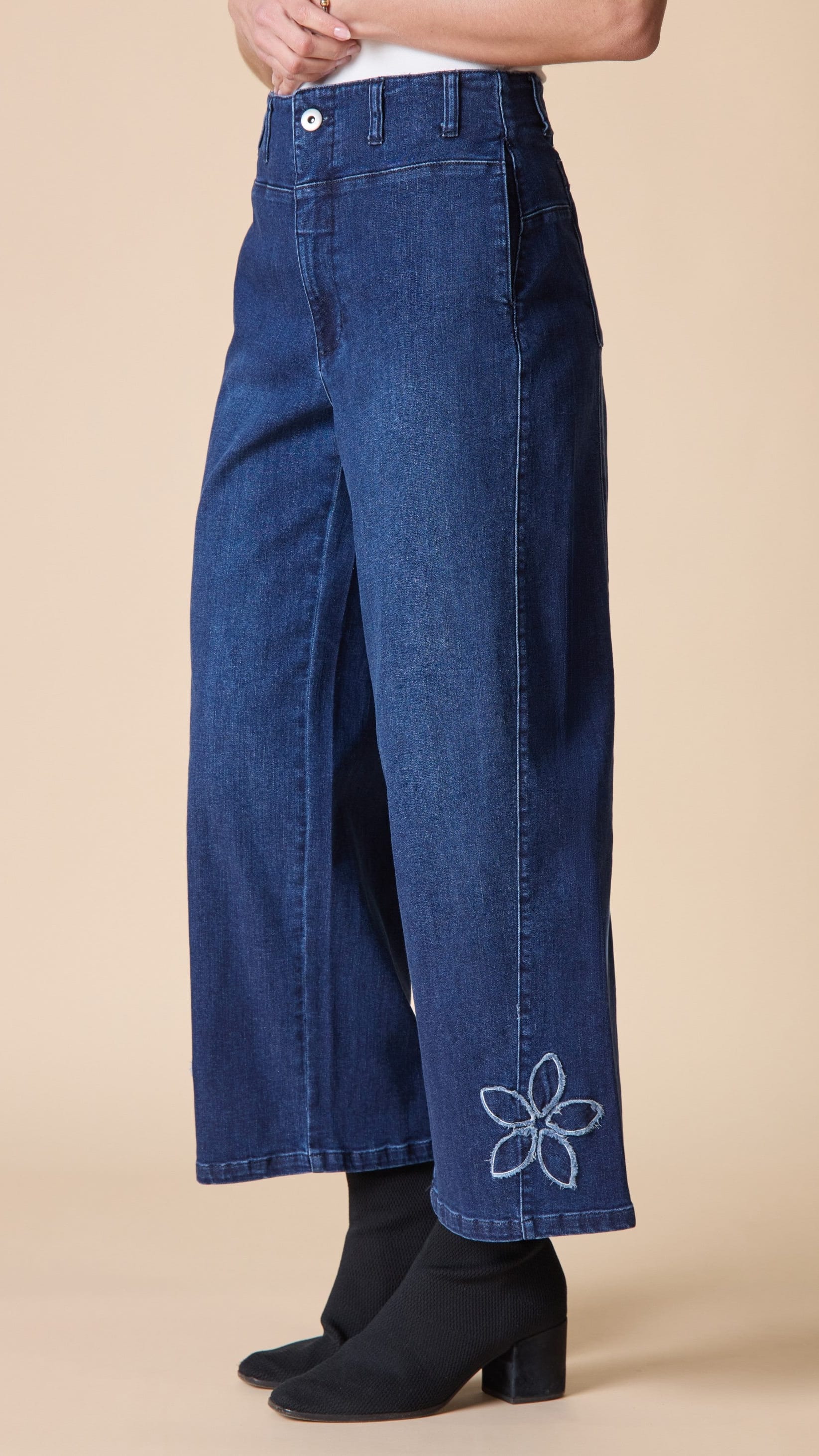 A person wearing dark blue wide-leg cropped jeans with a white embroidered flower detail near the ankle, paired with black ankle boots against a solid beige studio background.