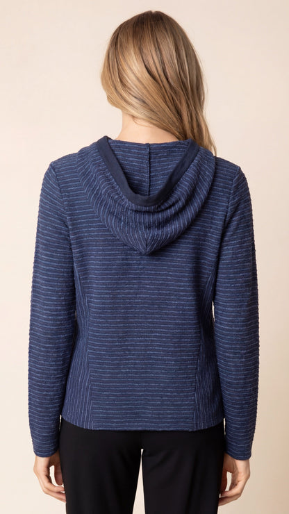 A woman viewed from the back wearing a navy blue hooded sweater with fine horizontal stripes and black pants, set against a plain beige studio background.