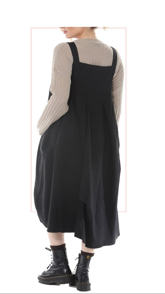 A model shown from the back wearing a black pinafore-style midi dress over a beige ribbed knit sweater, paired with black combat boots, on a white background with a thin pink rectangular outline.