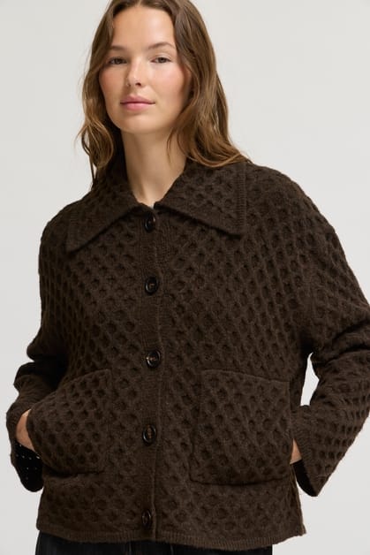 A woman wearing a dark brown textured knit button-up jacket with a wide collar and front pockets, photographed against a plain light gray studio background.