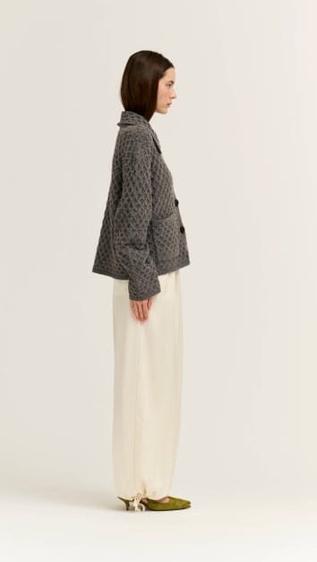 A woman stands in profile wearing a dark gray patterned quilted jacket, cream wide-leg trousers, and olive green shoes against a plain off-white studio background.
