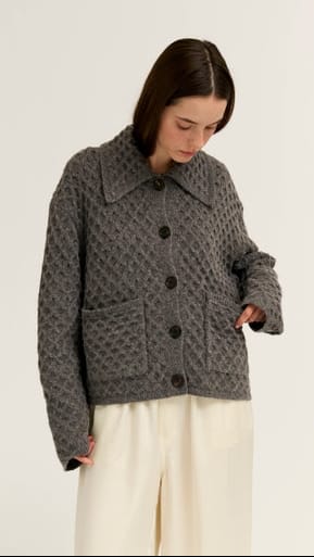 A woman wearing a dark gray textured knit cardigan with a collar and front buttons, paired with cream wide-leg trousers, photographed against a plain light background.