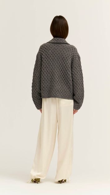 A model shown from the back wearing a dark gray textured knit cardigan and cream wide-leg trousers, standing against a plain off-white studio background.