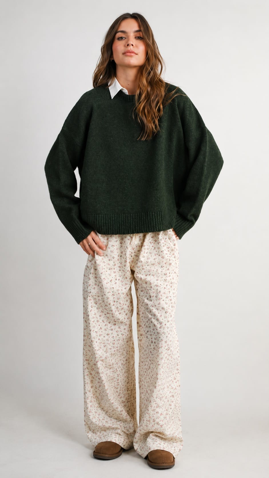 A woman stands against a light gray studio background wearing an oversized dark green knit sweater over a white collared shirt, loose cream patterned pants, and brown slip-on shoes.