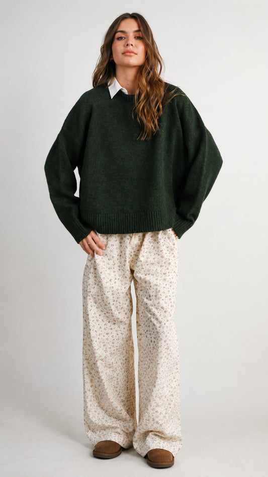 A woman stands against a light gray studio background wearing an oversized dark green knit sweater over a white collared shirt, loose cream patterned pants, and brown slip-on shoes.