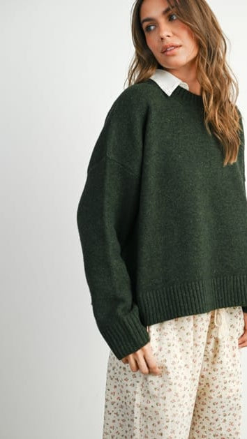 A woman wearing an oversized dark green knit sweater with a white pointed collar, paired with cream patterned trousers, photographed against a light gray studio background.
