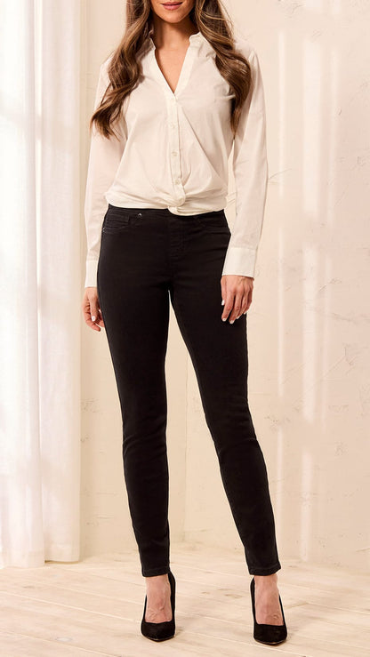 A woman modeling black skinny jeans with a cream wrap-style blouse and black heels, standing in a bright indoor setting with soft window light and curtains.