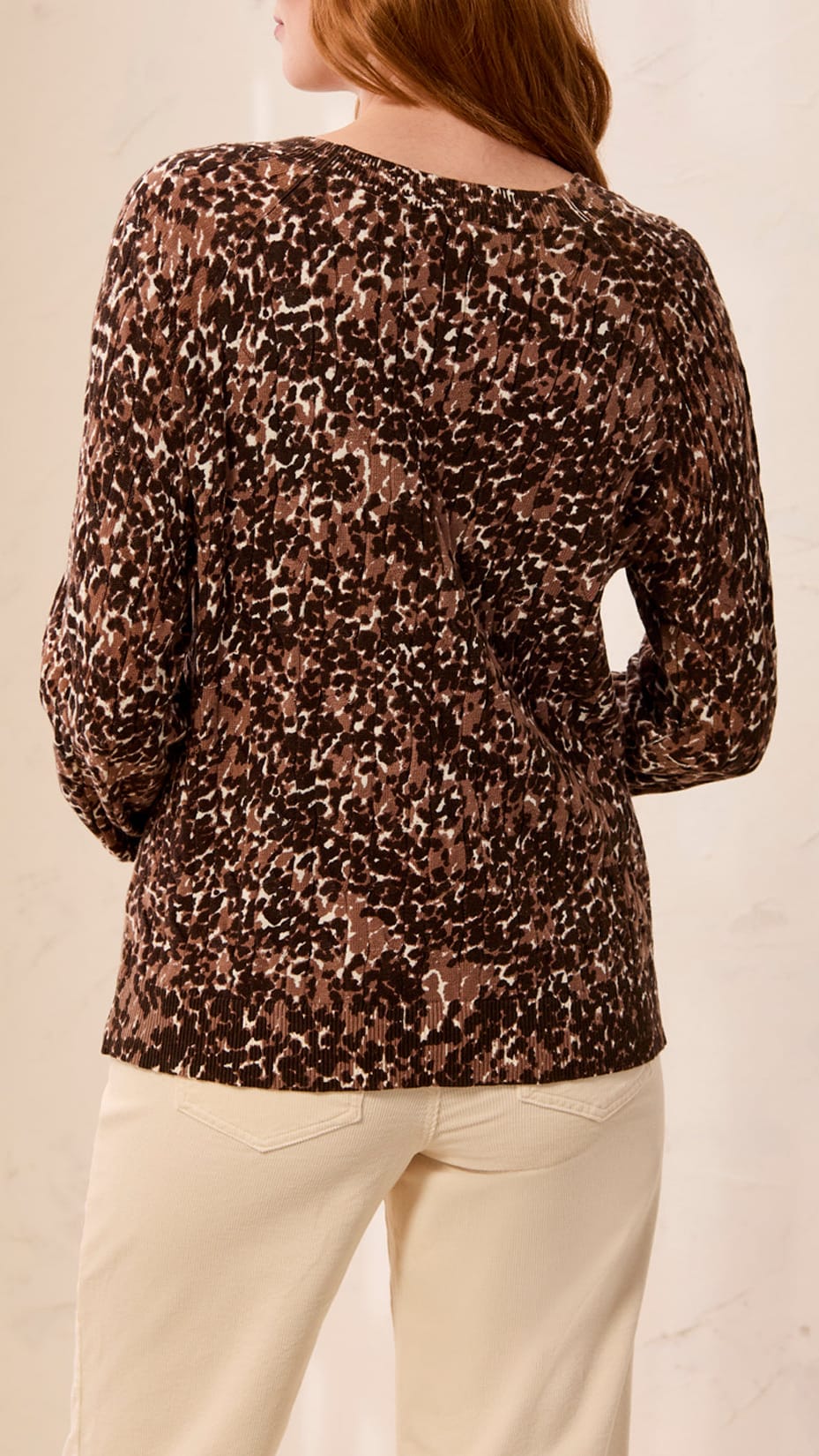 A woman shown from the back wearing a brown leopard-print long-sleeve top and cream pants against a light beige wall.