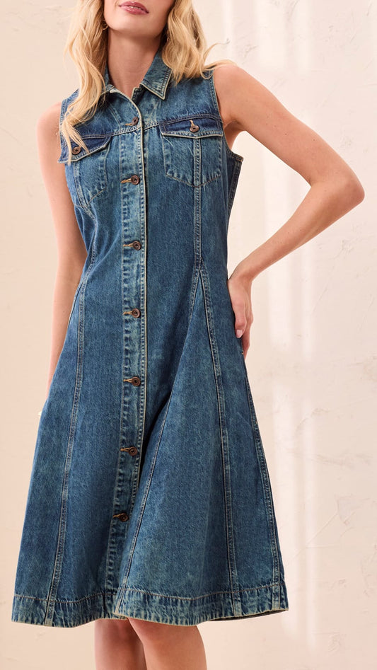 A woman wearing a sleeveless blue denim shirt dress with a button-front closure and chest pockets, standing against a light beige textured wall.
