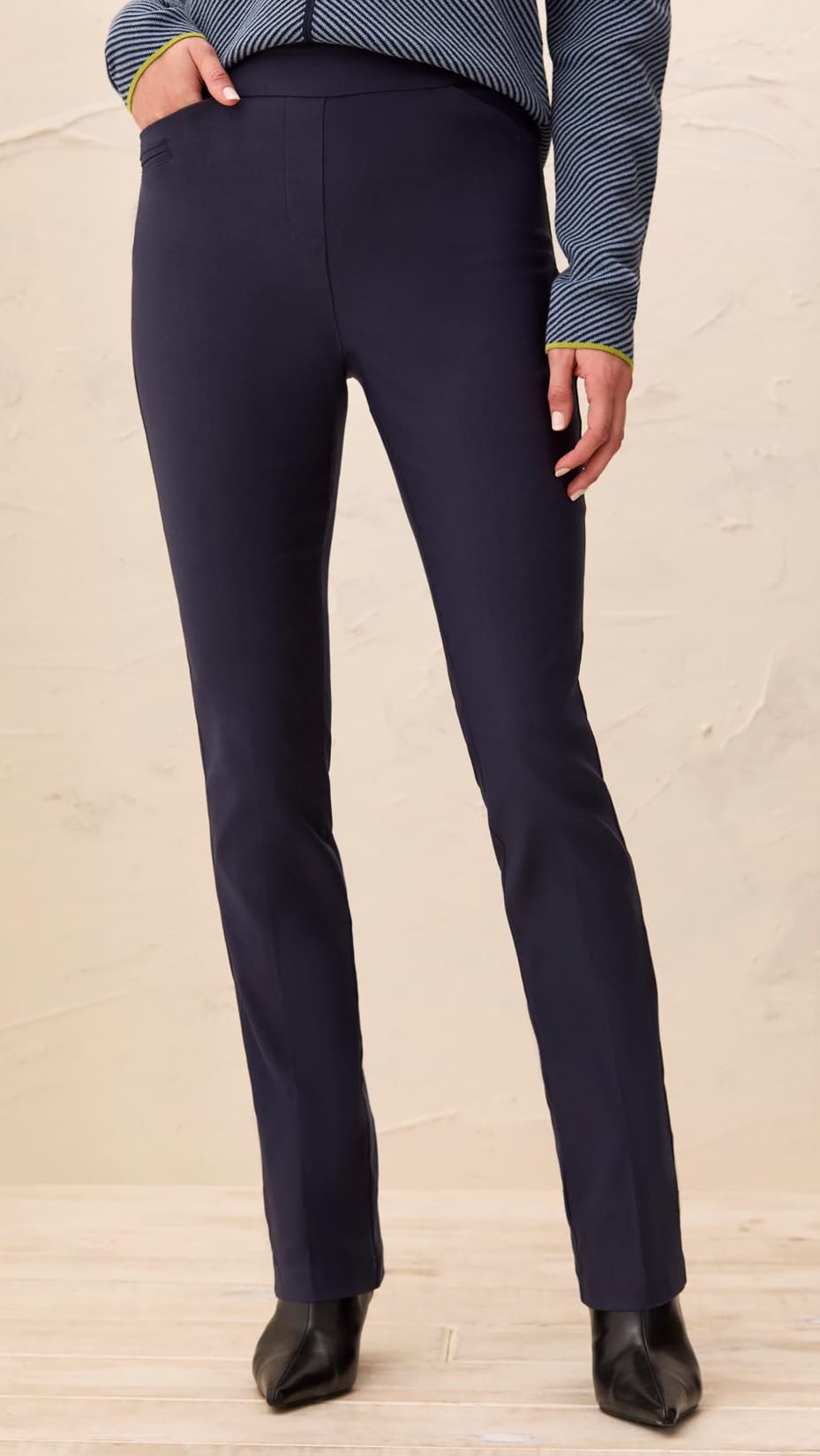 A person wearing dark navy straight-leg trousers, paired with a blue and white striped long-sleeve top and black pointed-toe heels, standing against a textured beige wall.