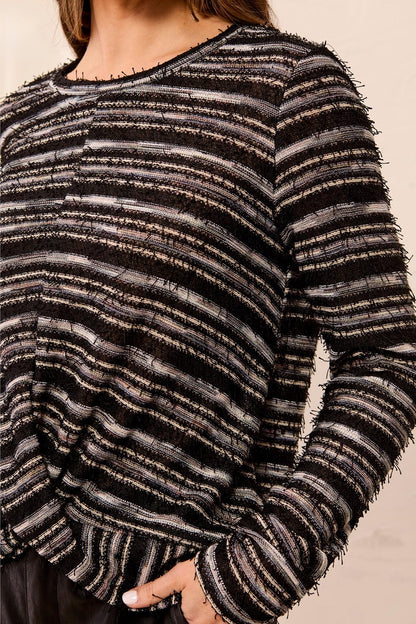 A close-up of a person wearing a long-sleeve knit top with horizontal black, gray, and white textured stripes and frayed edges.