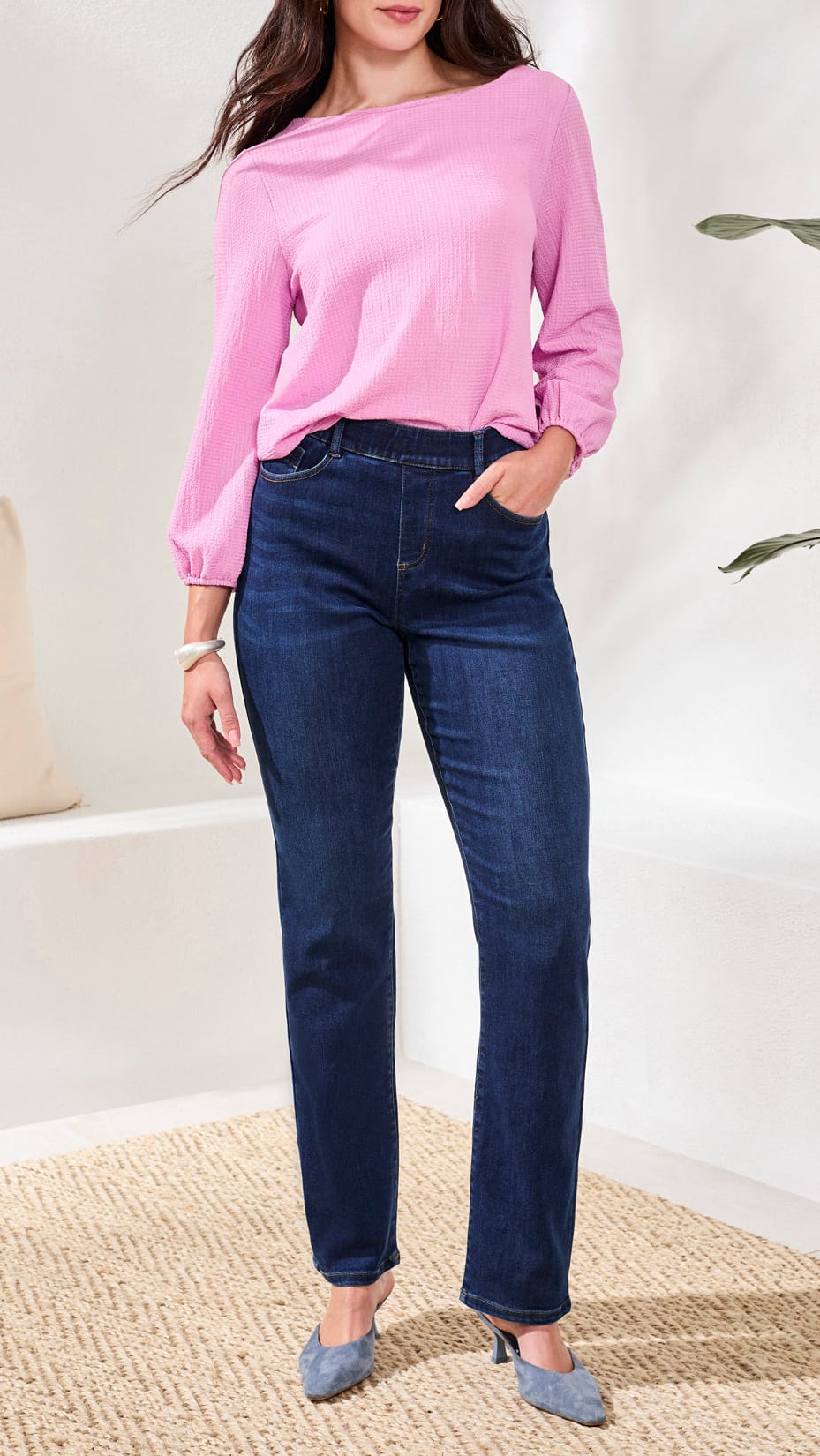 A woman modeling dark blue straight-leg jeans with a pink long-sleeve top and gray pointed-toe heels, standing in a bright indoor setting with a woven rug and a plant.