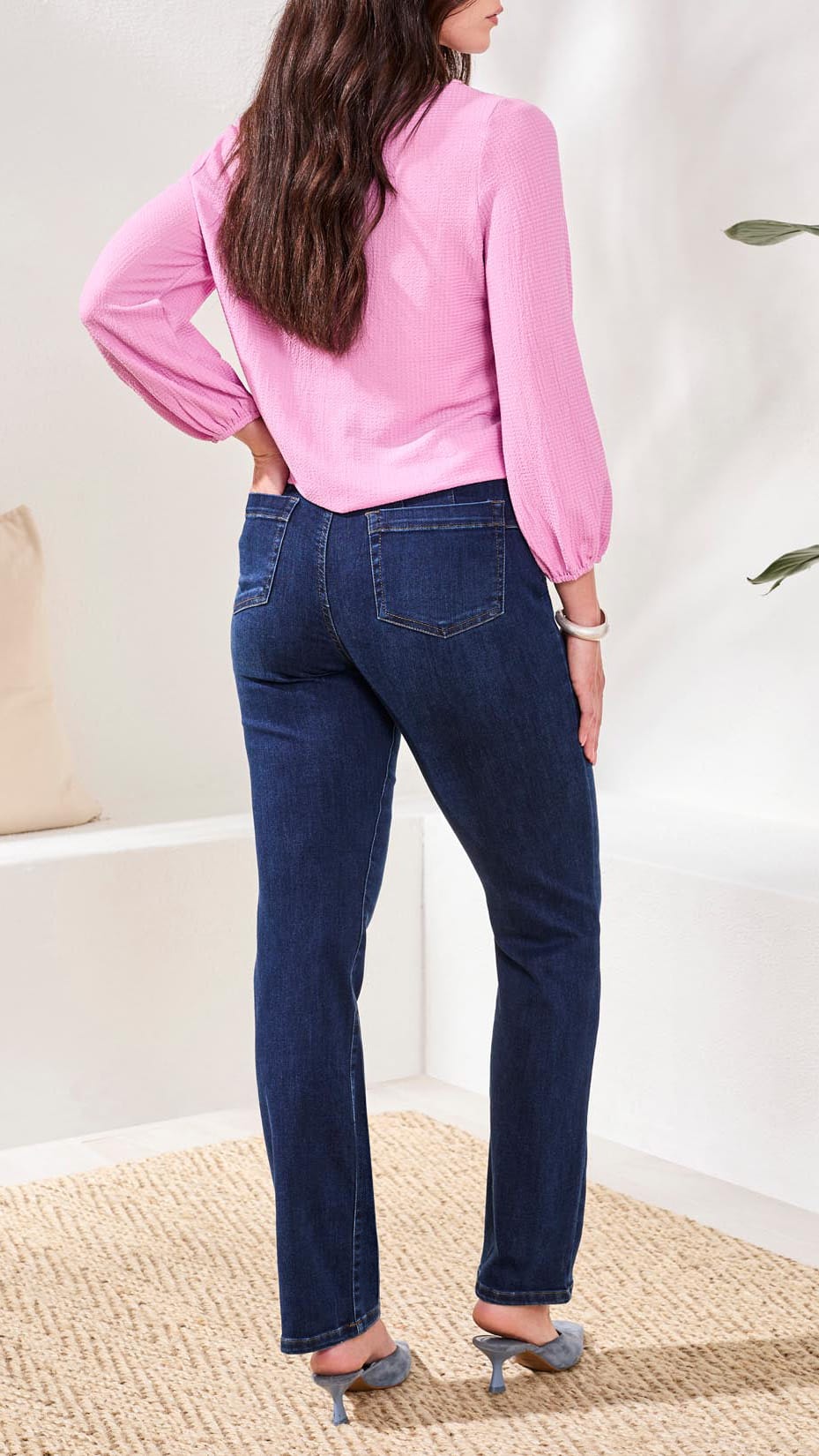 A woman shown from the back wearing dark blue skinny jeans and a pink long-sleeve blouse, standing on a woven rug in a bright indoor setting.