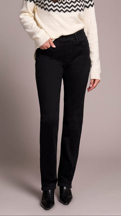 A woman modeling a cream knit sweater with a black and white geometric pattern across the chest and shoulders, paired with black jeans and black shoes, against a solid taupe studio background.
