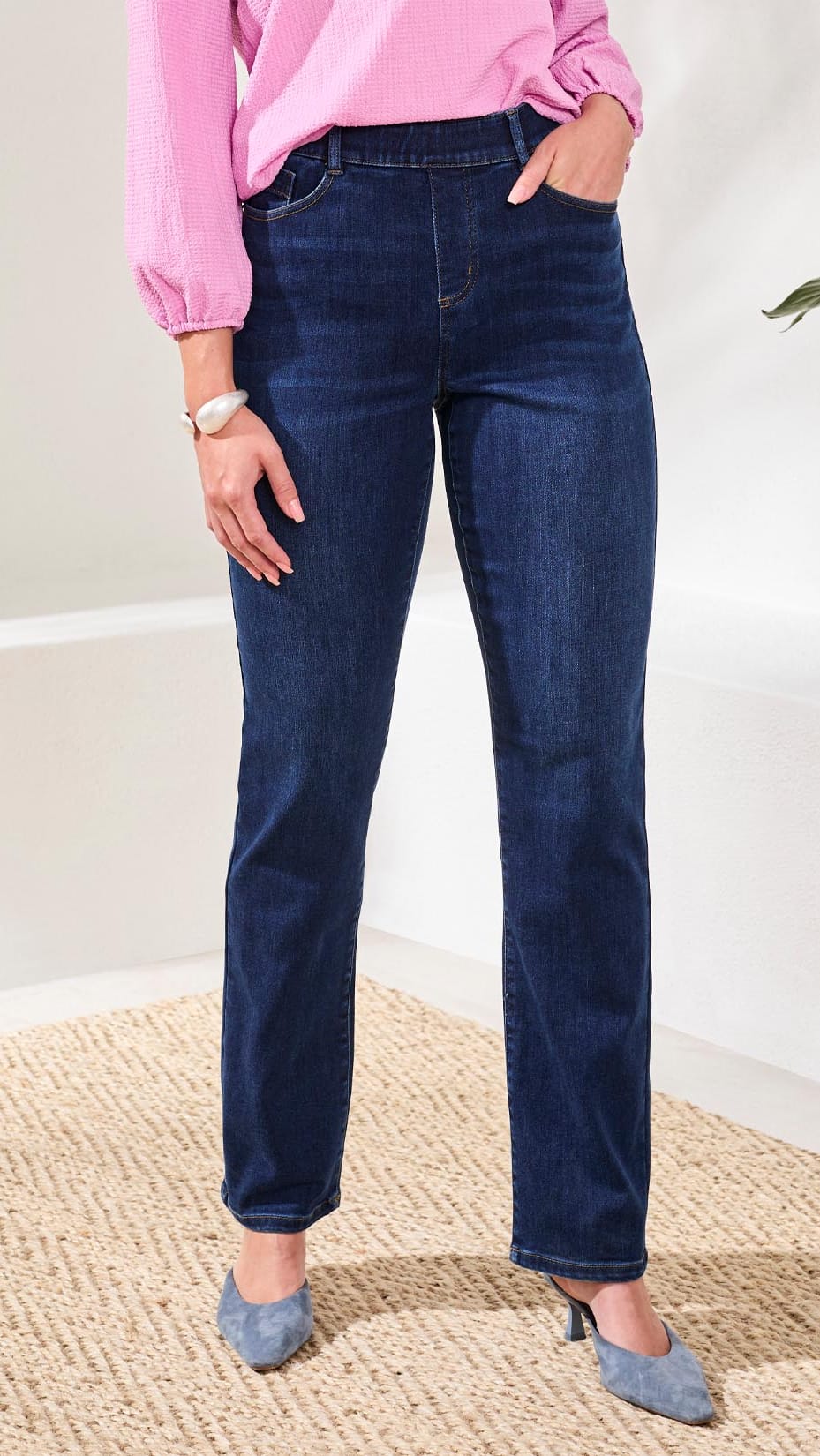 A person wearing dark blue straight-leg denim jeans, a pink long-sleeve top, and gray pointed-toe heels, standing on a woven rug in a bright indoor setting.