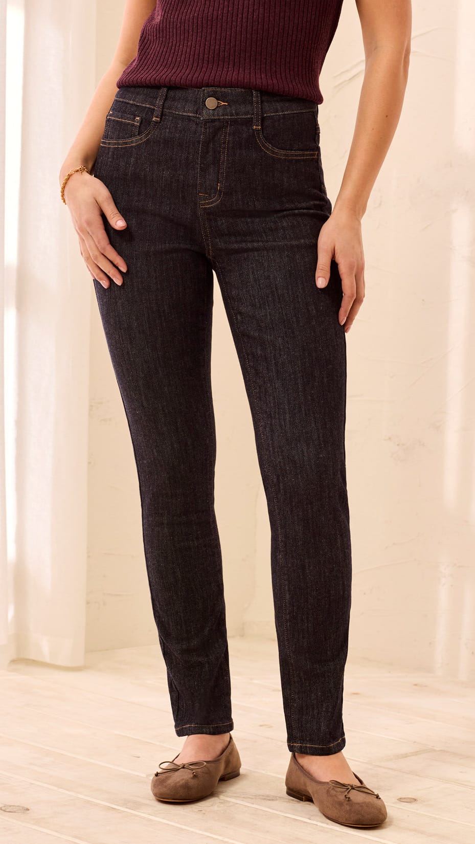 A person wearing dark indigo skinny jeans, a burgundy ribbed top, and brown flat shoes, standing in a brightly lit room with light-colored walls and wood flooring.